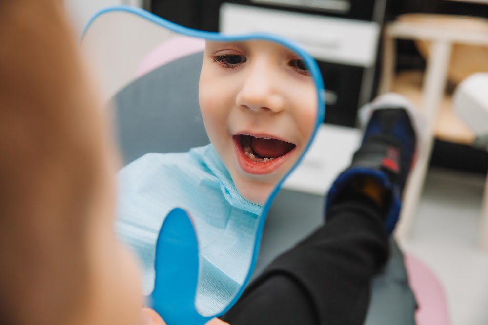boy-looks-mirror-with-toothy-smile-sitting-chair-with-dentist-dental-office A young child sitting in a dental chair, smiling during a routine pediatric dental check-up.